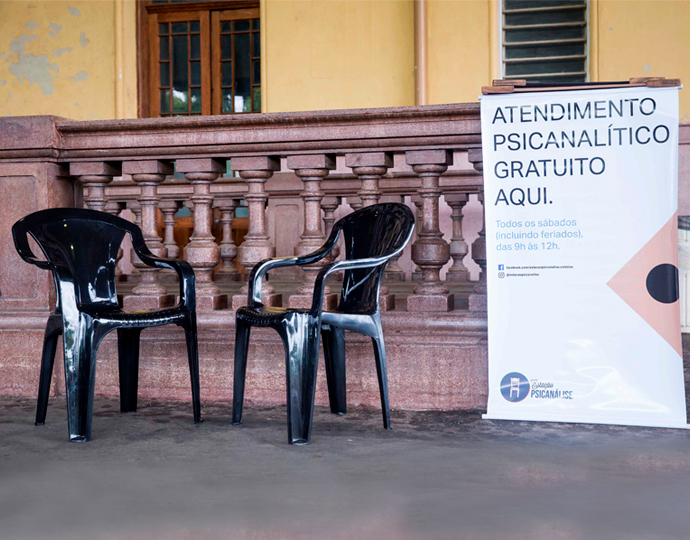 Foto mostra duas cadeiras pretas de plástico, dispostas em um ambiente da Estação Cultura de Campinas, ao lado de um banner com o texto "atendimento psicanalítico gratuito aqui". Clique enter para acessar